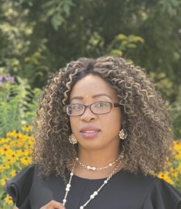 A woman with glasses and curly hair standing in front of flowers.